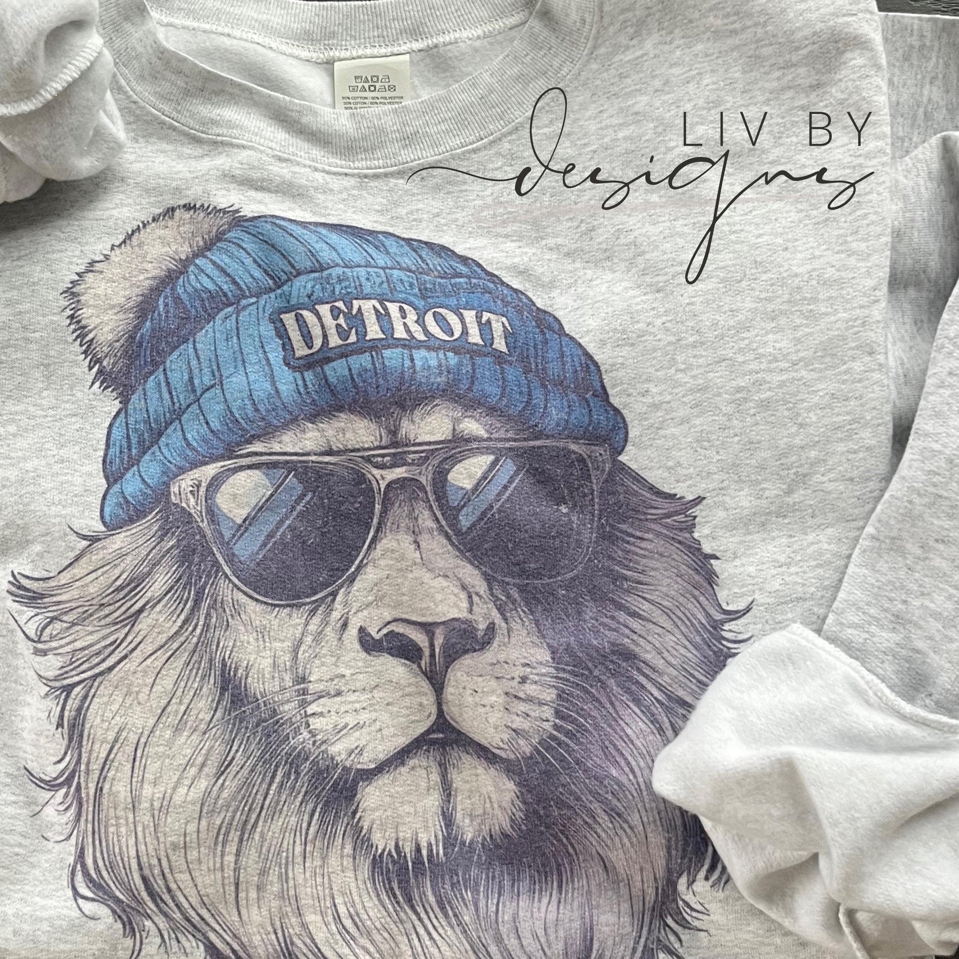 A gray sweatshirt with a graphic of a lion wearing a blue beanie with the word "DETROIT" on it and sunglasses. The lion has a white pom-pom on its beanie.