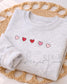A light gray sweatshirt with embroidered hearts in shades of pink and red. The design features five hearts in a row.