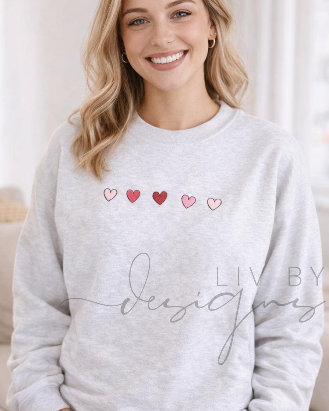 A light gray sweatshirt with five embroidered hearts in shades of pink and red. The text "LIV BY Design" is written in cursive script below the hearts. The sweatshirt is a casual, everyday garment.