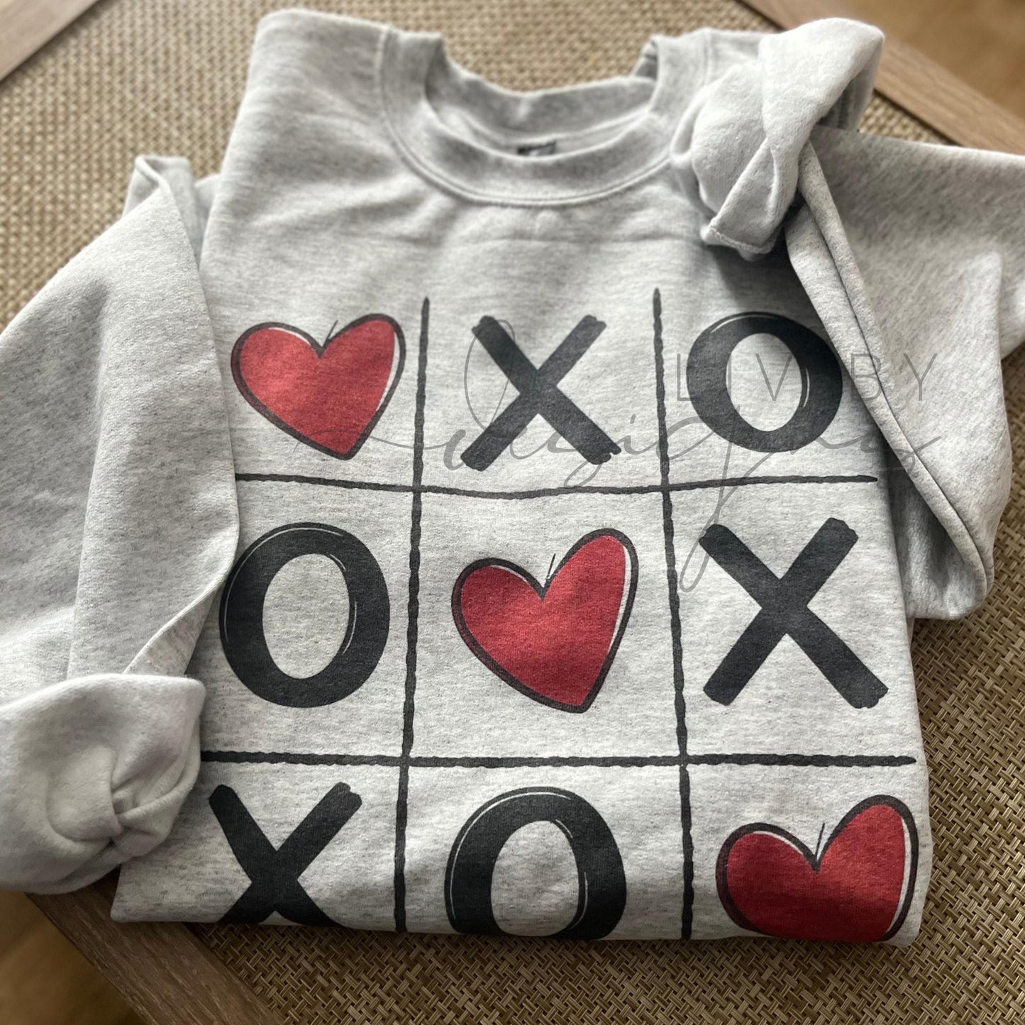 Ash grey Valentine’s Day crewneck sweatshirt featuring a tic tac toe design with Xs, Os, and red hearts, laid flat on a neutral background.