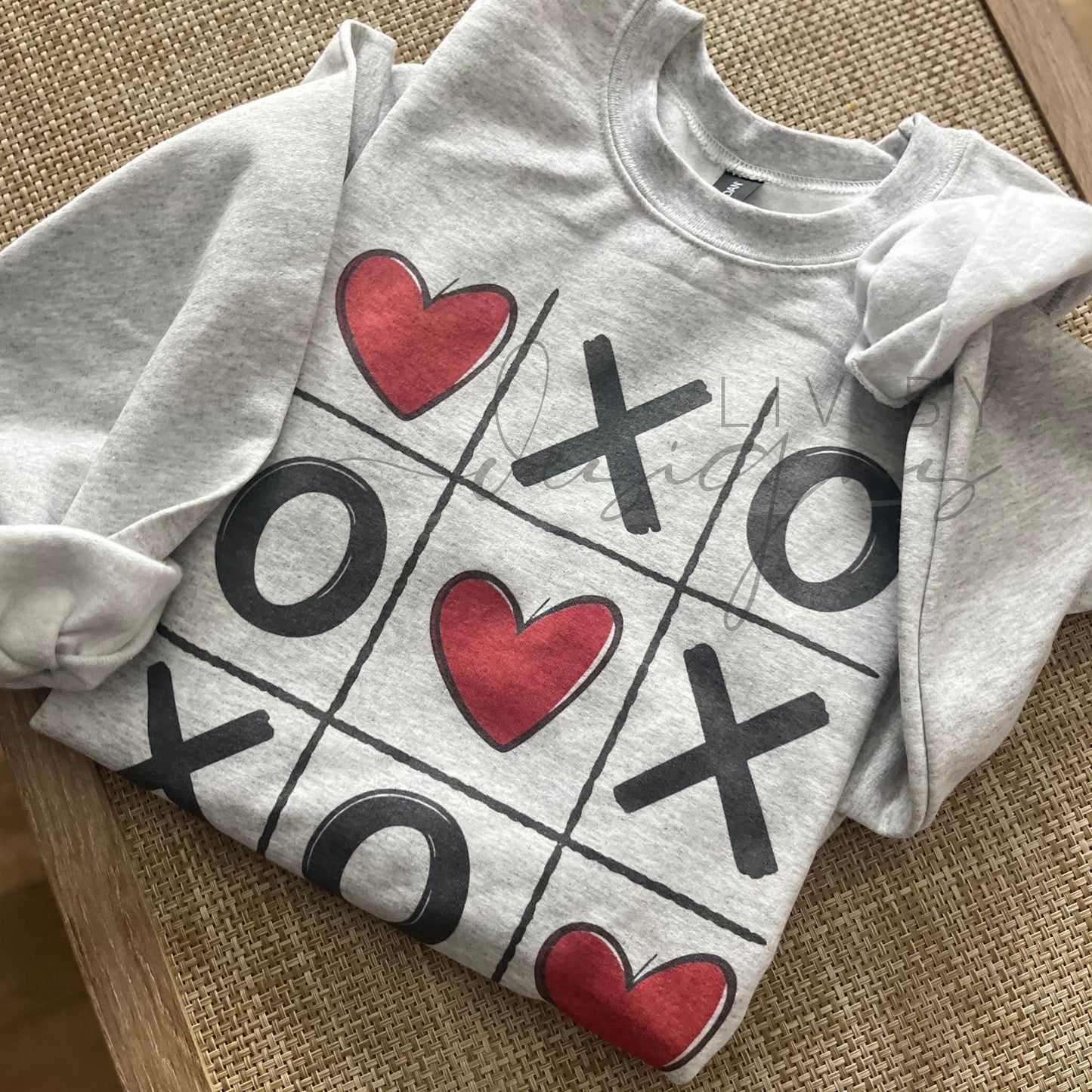 Ash grey Valentine’s Day crewneck sweatshirt featuring a tic tac toe design with Xs, Os, and red hearts, laid flat on a neutral background.