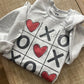 Ash grey Valentine’s Day crewneck sweatshirt featuring a tic tac toe design with Xs, Os, and red hearts, laid flat on a neutral background. (etsy image id: 7554599376)